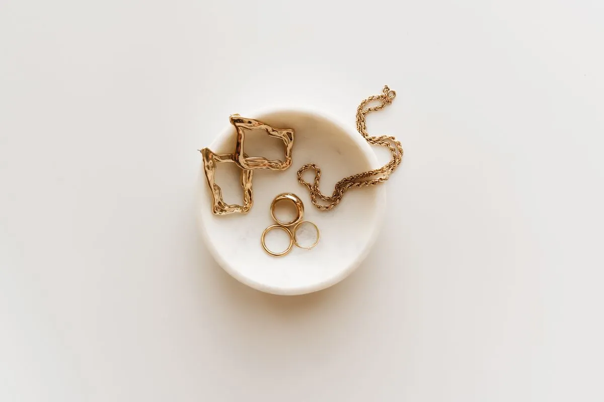 A gold chain, hoop earrings and three plain rings arranged in a small marble dish.