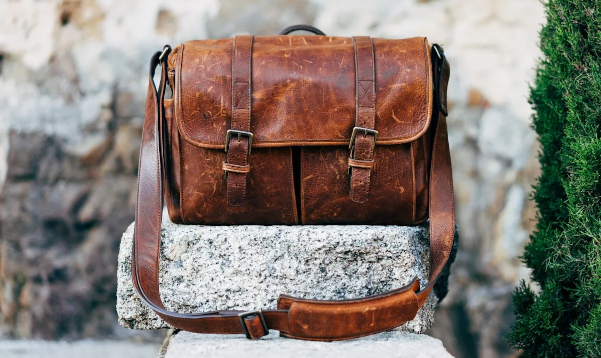 A worn brown leather satchel with brass buckles, standing on a stone wall.